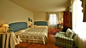 A hotel room with two single beds, a wooden floor, a green armchair, a small table with a lamp, and a large wooden cabinet. - Albergo Cannon D'Oro (Cocconato)