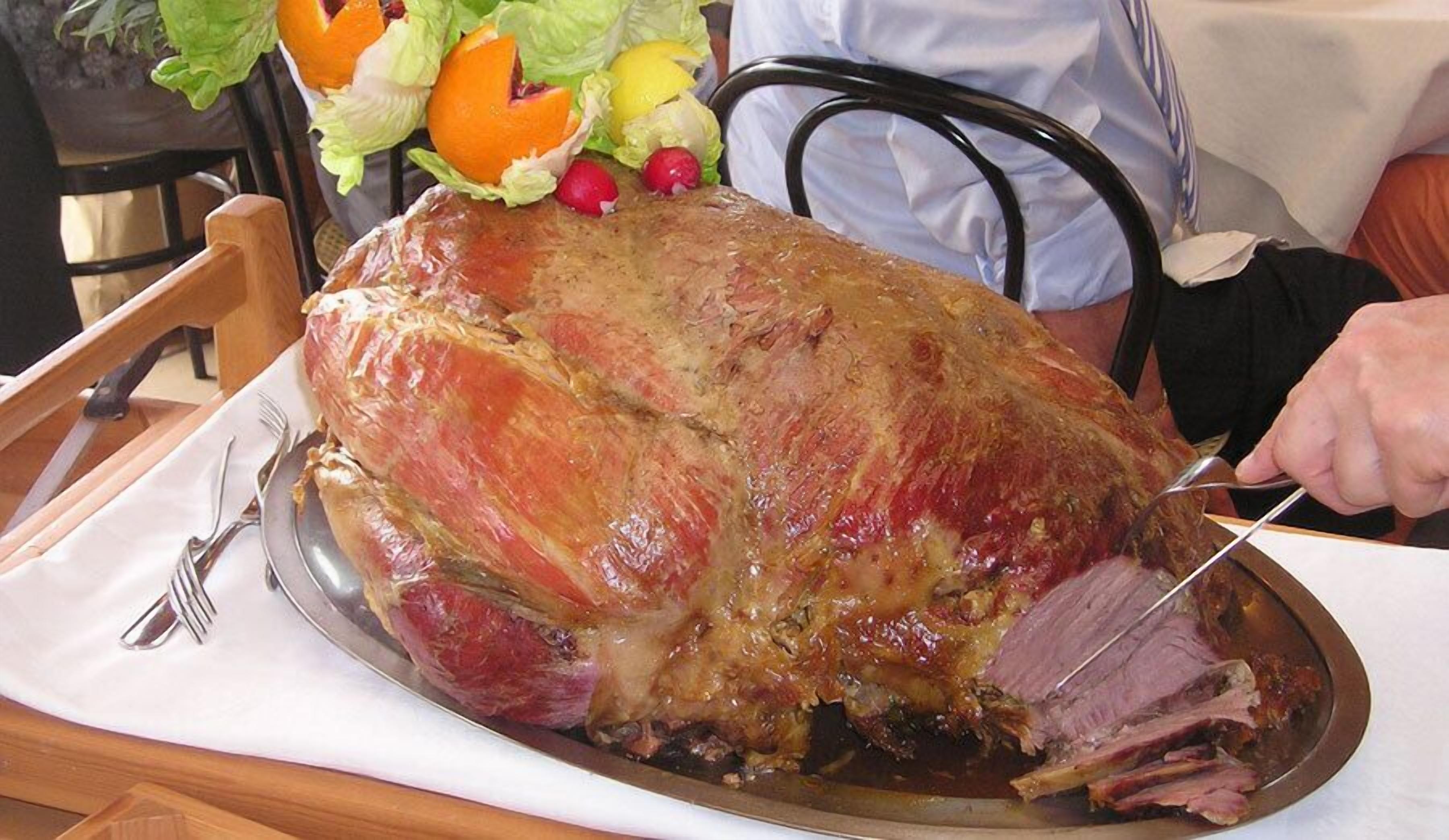 A large roasted ham on a platter with a carving fork and knife, surrounded by vegetables.