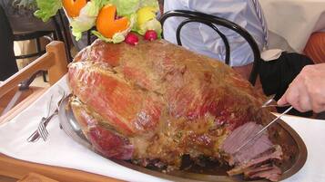 A large roasted ham on a platter with a carving fork and knife, surrounded by vegetables.