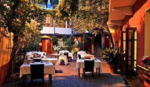 An outdoor dining area with tables and chairs, surrounded by lush greenery and a building with a tiled roof. - Albergo Cannon D'Oro (Cocconato)