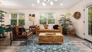 A spacious living room with a brown leather sofa, a wooden coffee table, and a large window with a view of greenery. - Hot Tub | New Build | Sauna | Game Room | Walk Downtown | Theater | Arcade (Ann Arbor)