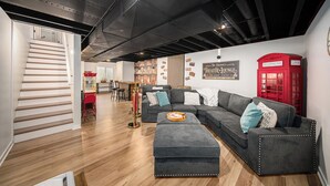A modern living room with a sectional sofa, a red telephone booth, and a staircase leading to another area. - Hot Tub | New Build | Sauna | Game Room | Walk Downtown | Theater | Arcade (Ann Arbor)