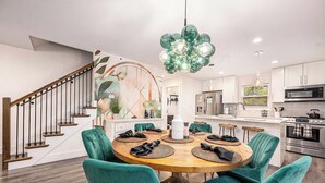 A modern kitchen with white cabinets, a wooden dining table with teal chairs, and a unique green chandelier. - Hot Tub | New Build | Sauna | Game Room | Walk Downtown | Theater | Arcade (Ann Arbor)