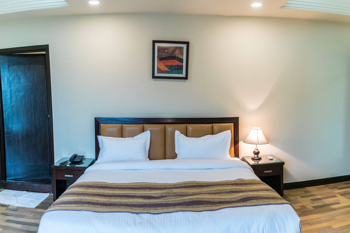 A hotel room with a large bed, a nightstand with a lamp, a framed picture on the wall, and a door.