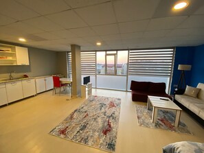 A modern living room with a sofa, a coffee table, and a TV. There is a kitchen area with cabinets and a dining table. - Su Residence (Trabzon)