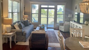 A spacious living room with a grey sofa, a wicker chair, a dining table with chairs, a ceiling fan, and large windows with a view of the outdoors.