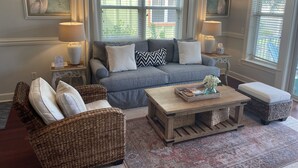 A living room with a grey sofa, wicker chairs, a coffee table, and two windows with blinds.