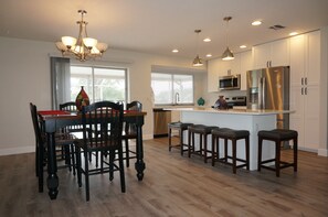 A modern kitchen with a dining area, featuring a chandelier, pendant lights, a central island with stools, and stainless steel appliances. - Living on the Green- Pool home with sunset views (Fort Myers)