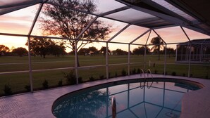 Una piscina interior con cerramiento de vidrio, rodeada de césped y árboles, durante el atardecer.