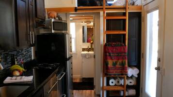 A compact kitchen with a microwave, stove, and cabinets. There is a small dining area with a table and chairs. A loft bed is visible in the background.