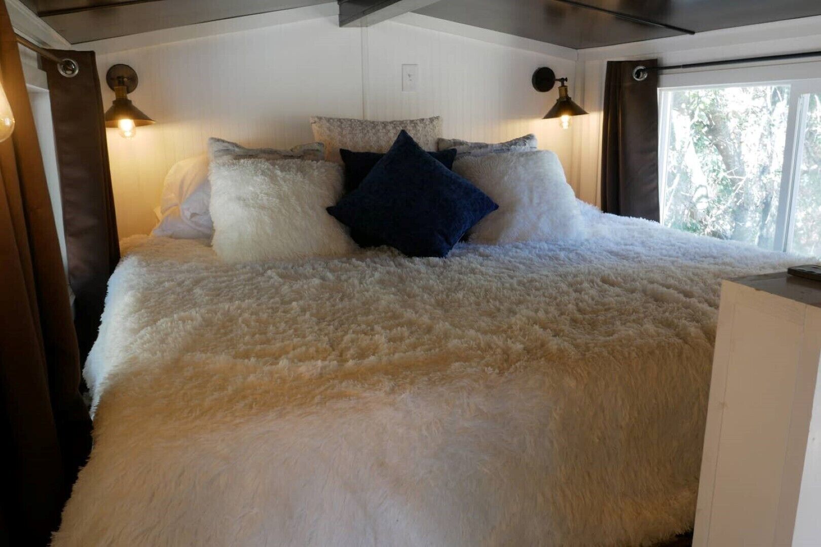 A bed with a fluffy white cover, two wall-mounted lamps, and a window with a view of trees.