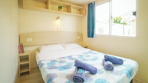 Una cama bien hecha con una colcha estampada y toallas enrolladas, una mesita de noche con un libro y una lámpara, y una ventana con cortina.