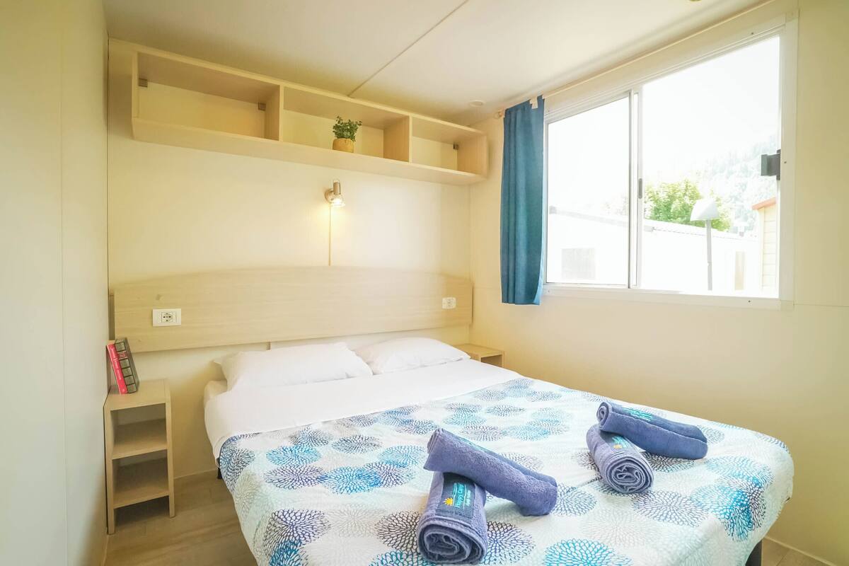 Een keurig opgemaakt bed met een beddengoed met patroon en opgerolde handdoeken, een nachtkastje met een boek en een lamp, en een raam met een gordijn.