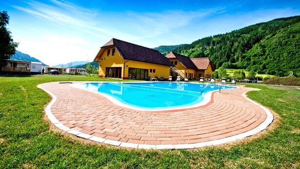 Una piscina con borde de ladrillo, rodeada de césped y un edificio amarillo con techo rojo.