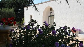 A white building with an arched doorway and a small window. There are potted plants with red and purple flowers in front of the building. - Authentic Trullo & Villa with Pool – Perfect for Families & Groups (Ceglie Messapica)
