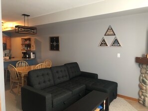 Uma sala de estar com sofá modular, uma área de jantar com cadeiras de madeira, uma cozinha com armários de madeira e uma parede de pedra.