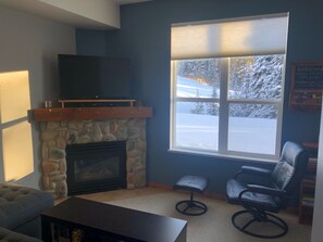 Uma sala de estar com lareira de pedra, uma televisão de tela plana montada acima dela, uma janela grande com vista para uma paisagem nevada, uma poltrona de couro e um pequeno banquinho.