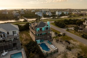 A multi-story beachfront property with a swimming pool, outdoor seating, and a view of the water and surrounding houses.
