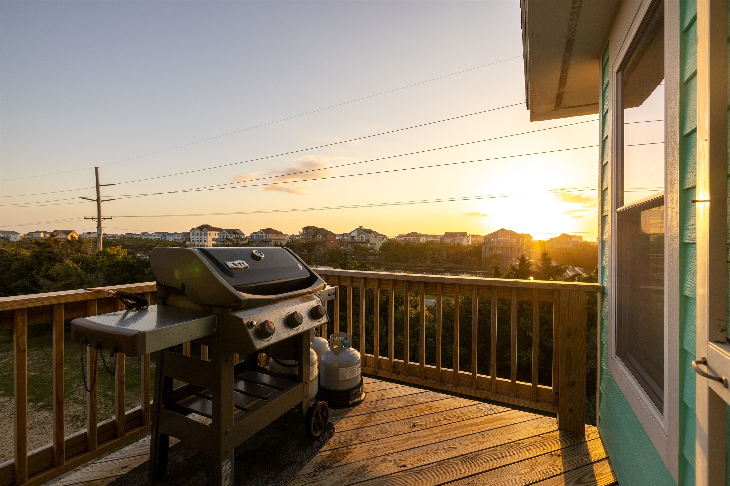 Une terrasse en bois équipée d’un barbecue et de bouteilles de propane, donnant sur un quartier résidentiel au coucher du soleil.