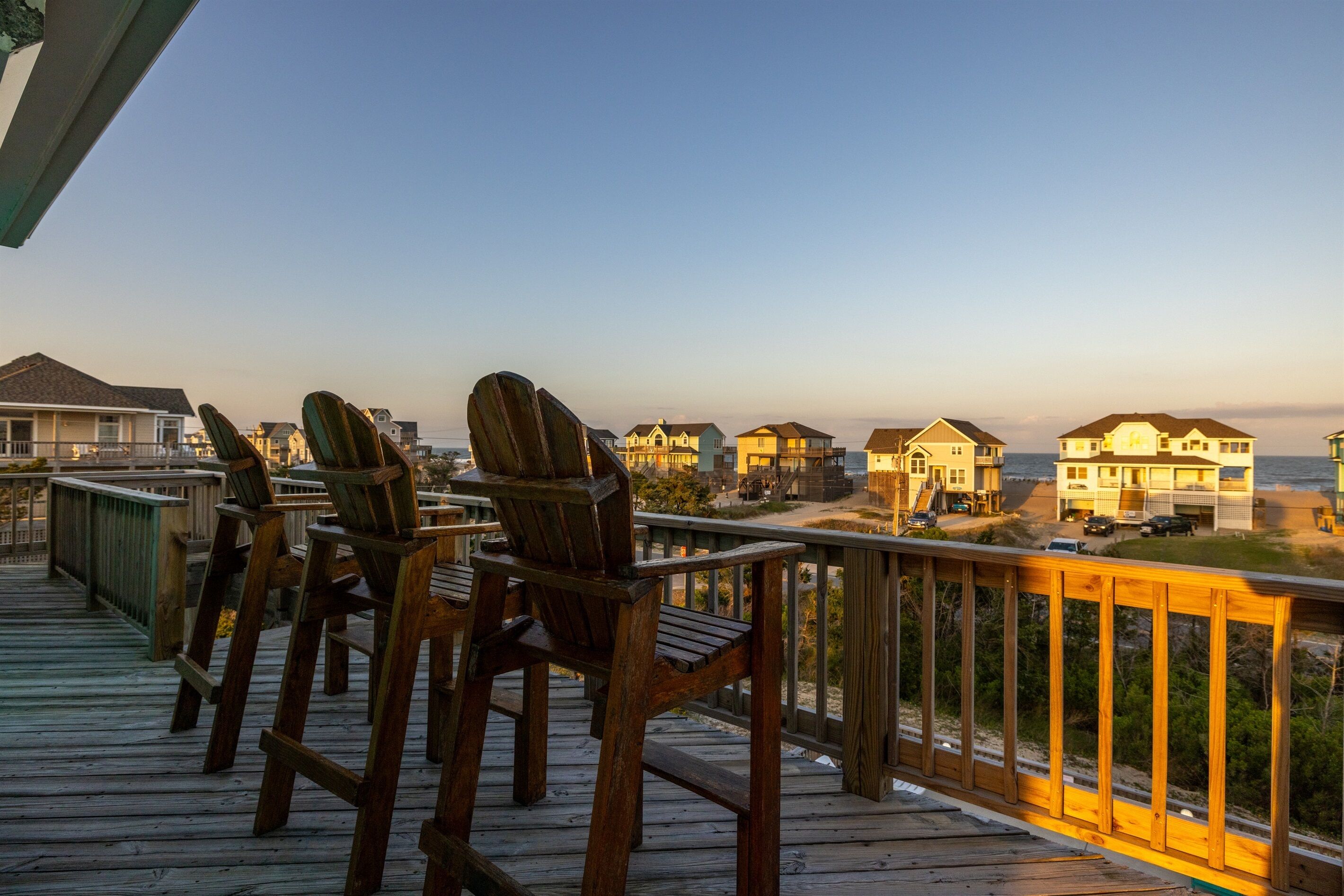 Une terrasse en bois avec des chaises, donnant sur une plage où l’on aperçoit des maisons au loin.