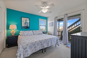 A bedroom with a teal wall, a bed with a floral bedspread, a ceiling fan, a nightstand with a lamp, and a view of a building through the window.