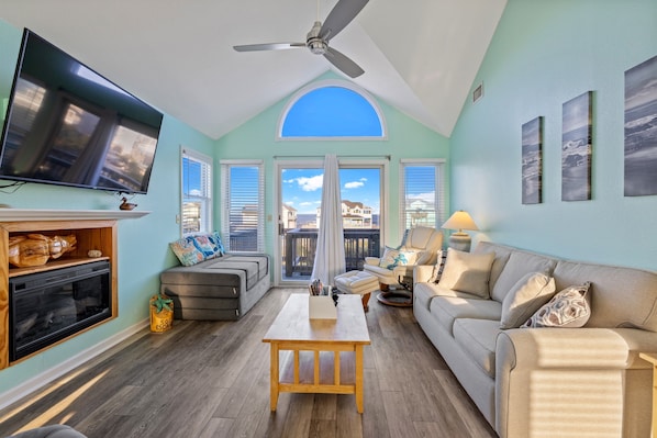 A living room with a TV, fireplace, sofa, and a view of houses through large windows.