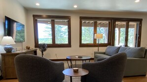 Ett vardagsrum med två grå fåtöljer, en grå soffa, ett litet runt bord och en lampa på ett litet sidobord i trä. Det finns två fönster med träramar och utsikt över träd utanför.