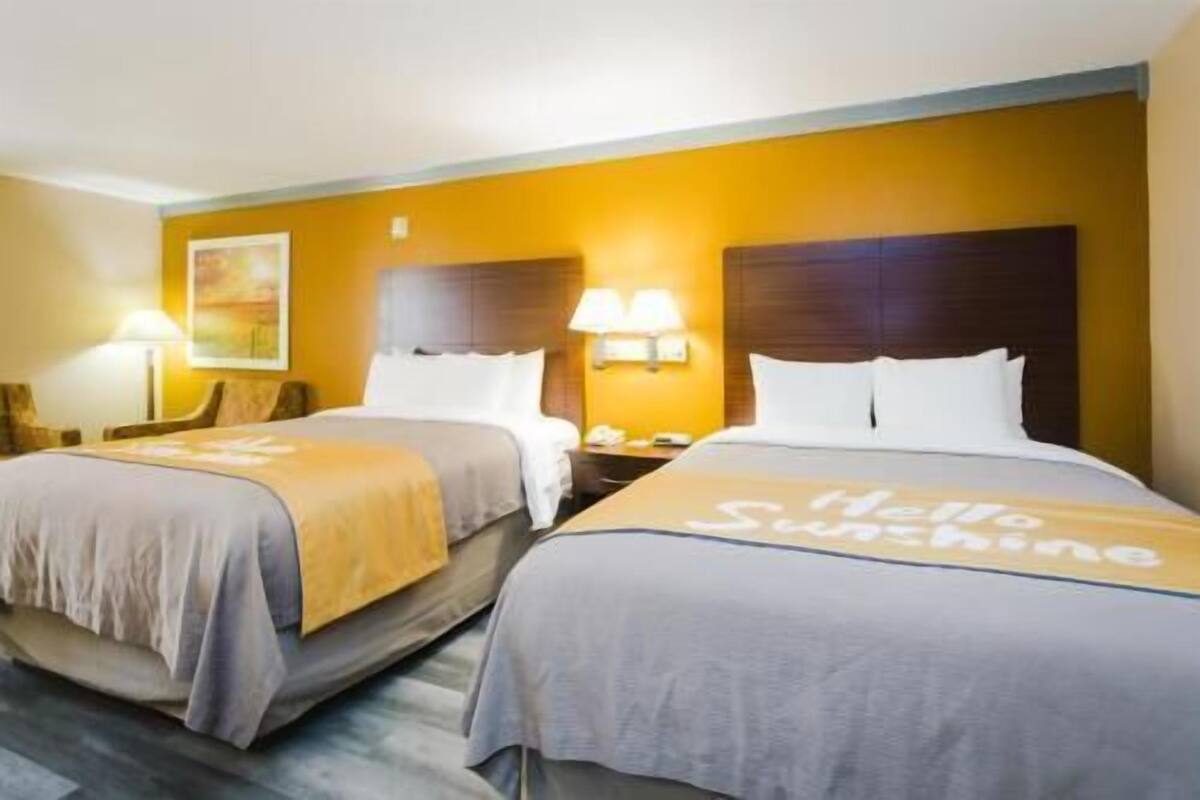 A hotel room with two beds, each with a headboard, bedside lamps, and a 'Hello Sunshine' bedspread.