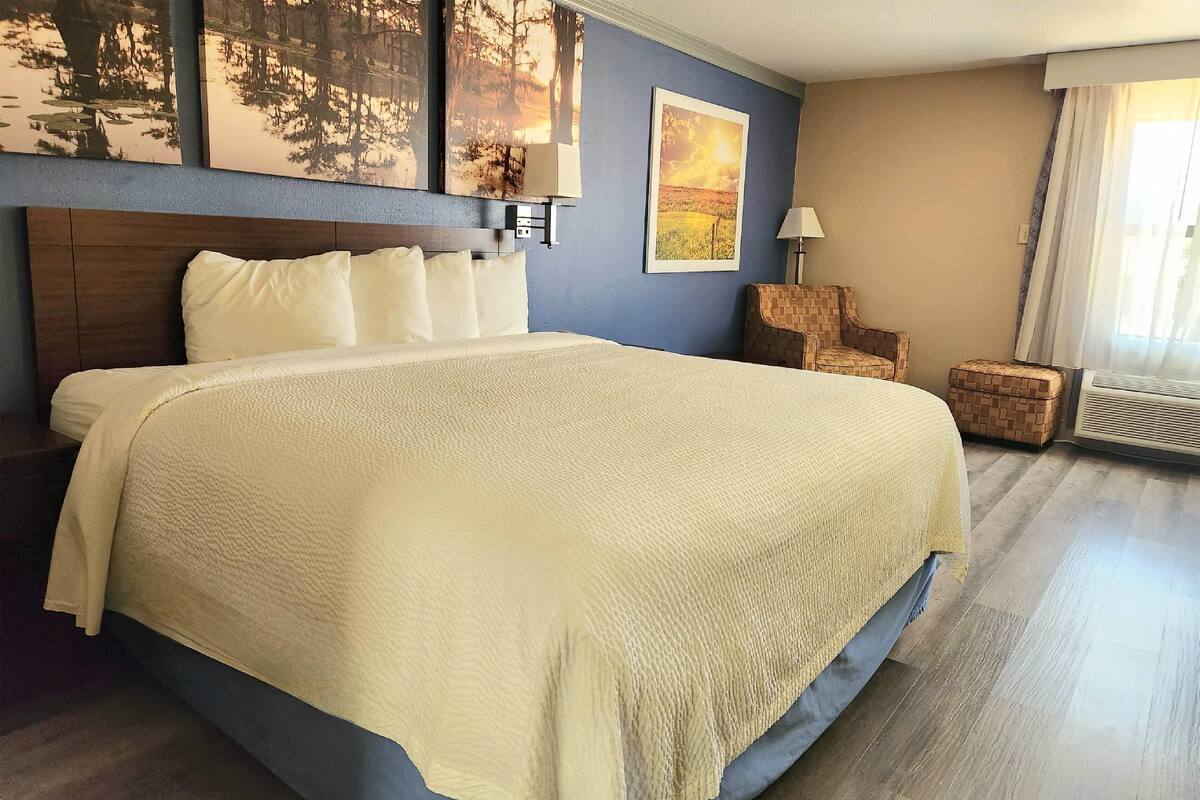 A hotel room with a large bed, a wooden headboard, a chair, a small table, and a window with curtains.