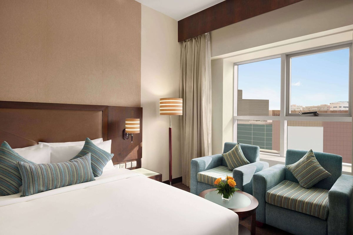 A hotel room with a large bed, two armchairs, a small table with flowers, and a window with a city view.