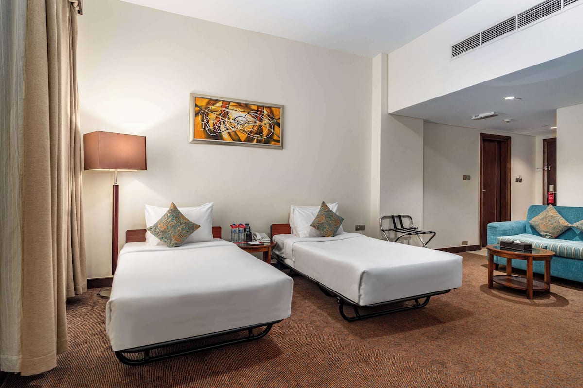 A hotel room with two single beds, a sofa, a lamp, and a painting on the wall.