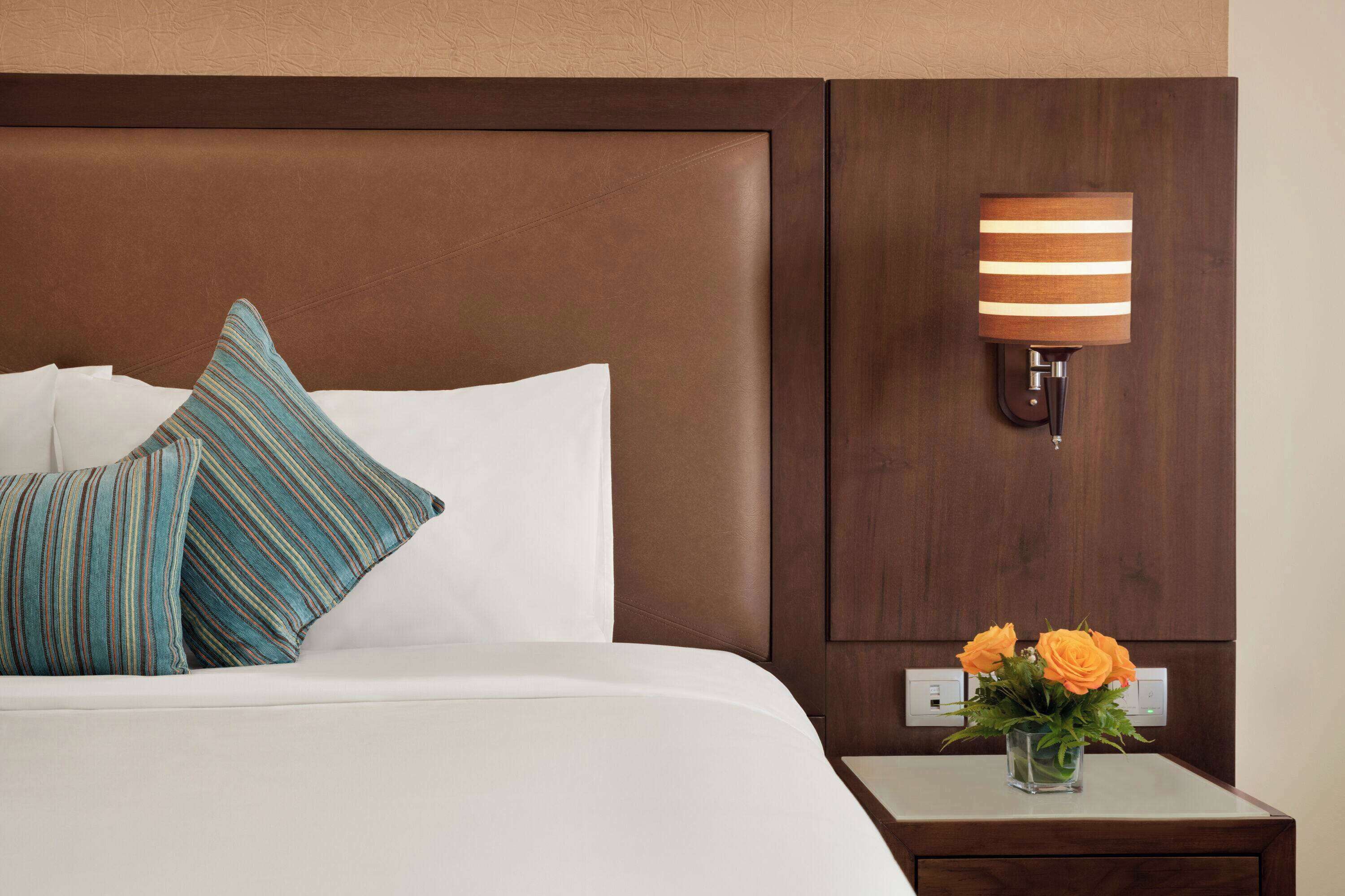 Una cama bien hecha con almohadas a rayas, una lámpara de pared y una mesita con un jarrón de flores.