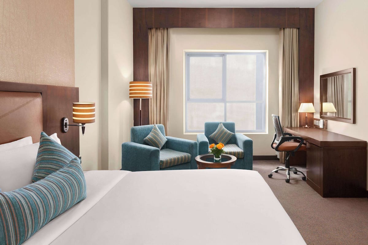 A hotel room with a large bed, two armchairs, a desk, a chair, and a window with curtains.