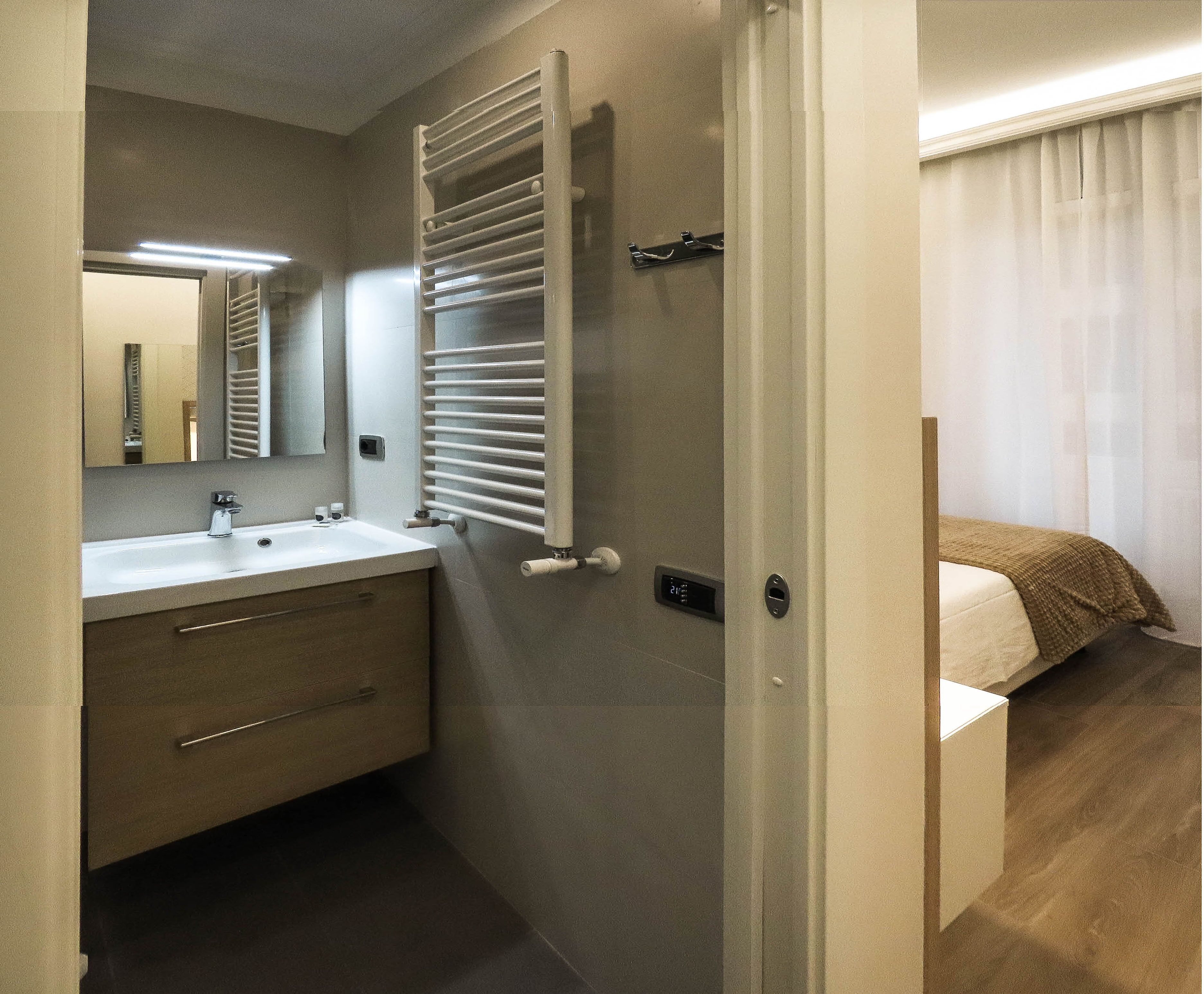 A modern bathroom with a sink, mirror, towel rack, and a door leading to a bedroom with a bed and curtains.