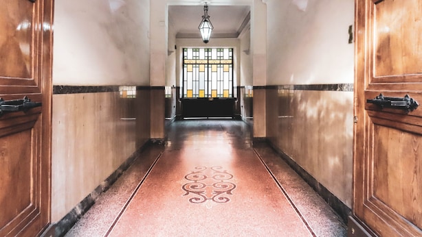 A hallway with wooden doors and a patterned floor leading to a room with a large window and a hanging light fixture. - My Maison 109 (Rome)