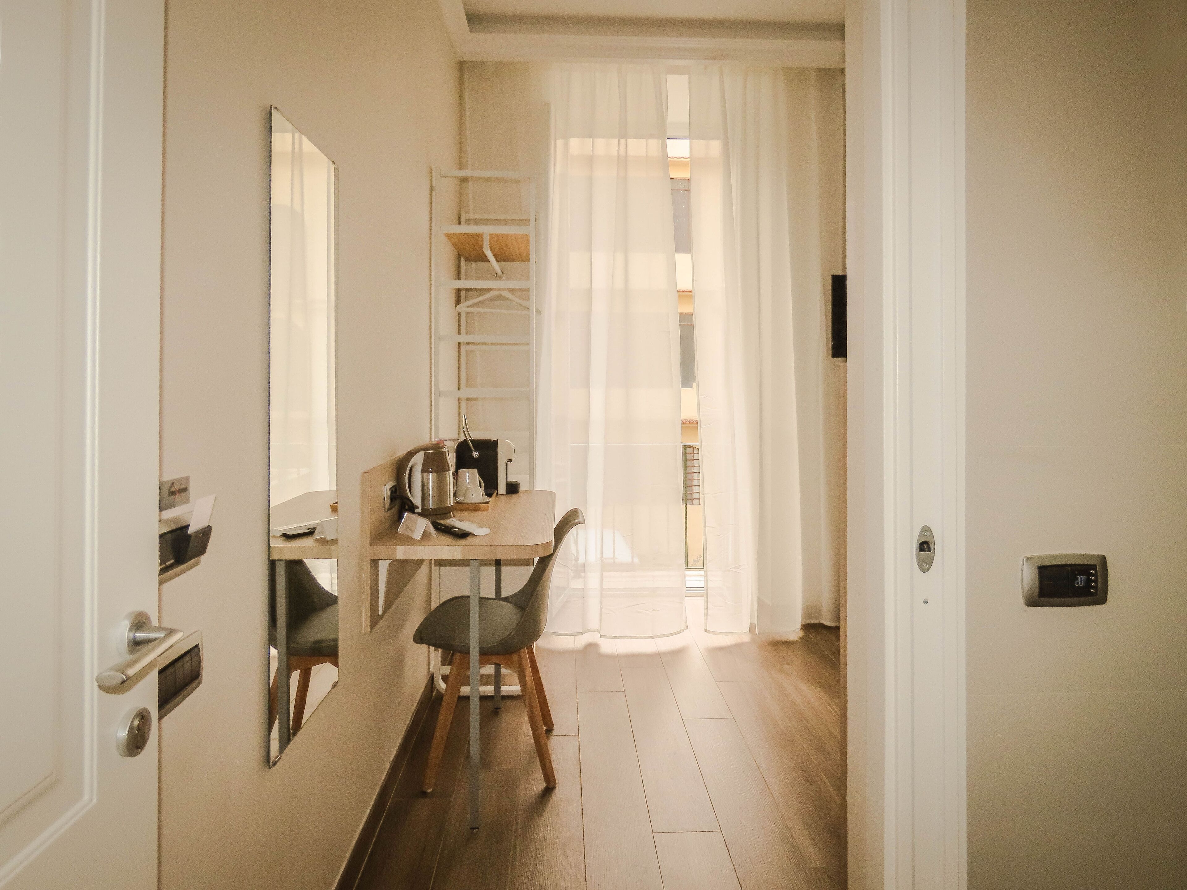 A small, bright room with a wooden table, a chair, a coffee maker, and a window with sheer curtains.