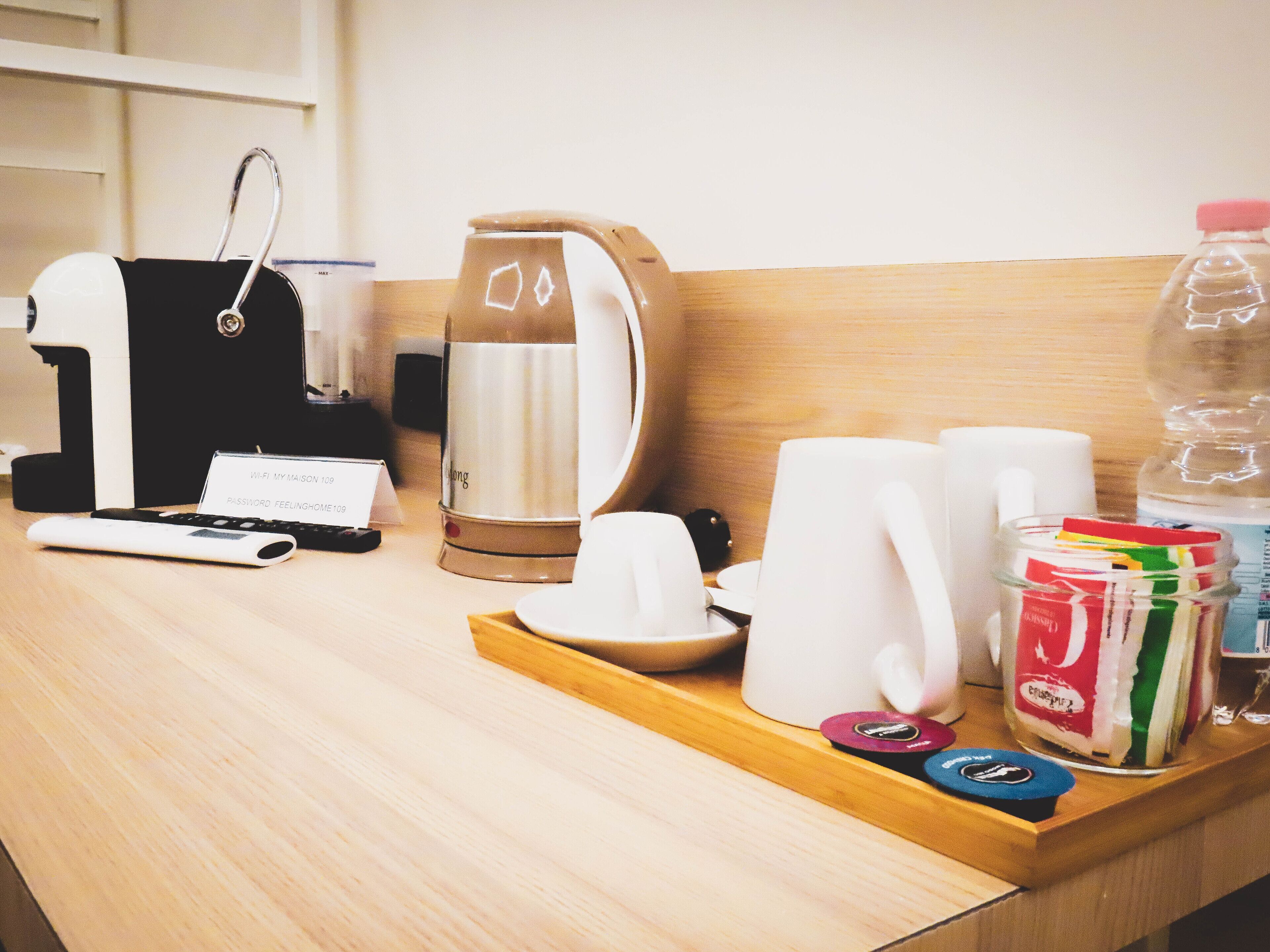 Un espace d’accueil dans une chambre d’hôtel, comprenant une machine à café, une bouilloire, un plateau avec des tasses et un pot de condiments, ainsi qu’un panneau indiquant la disponibilité du Wi-Fi et le mot de passe.