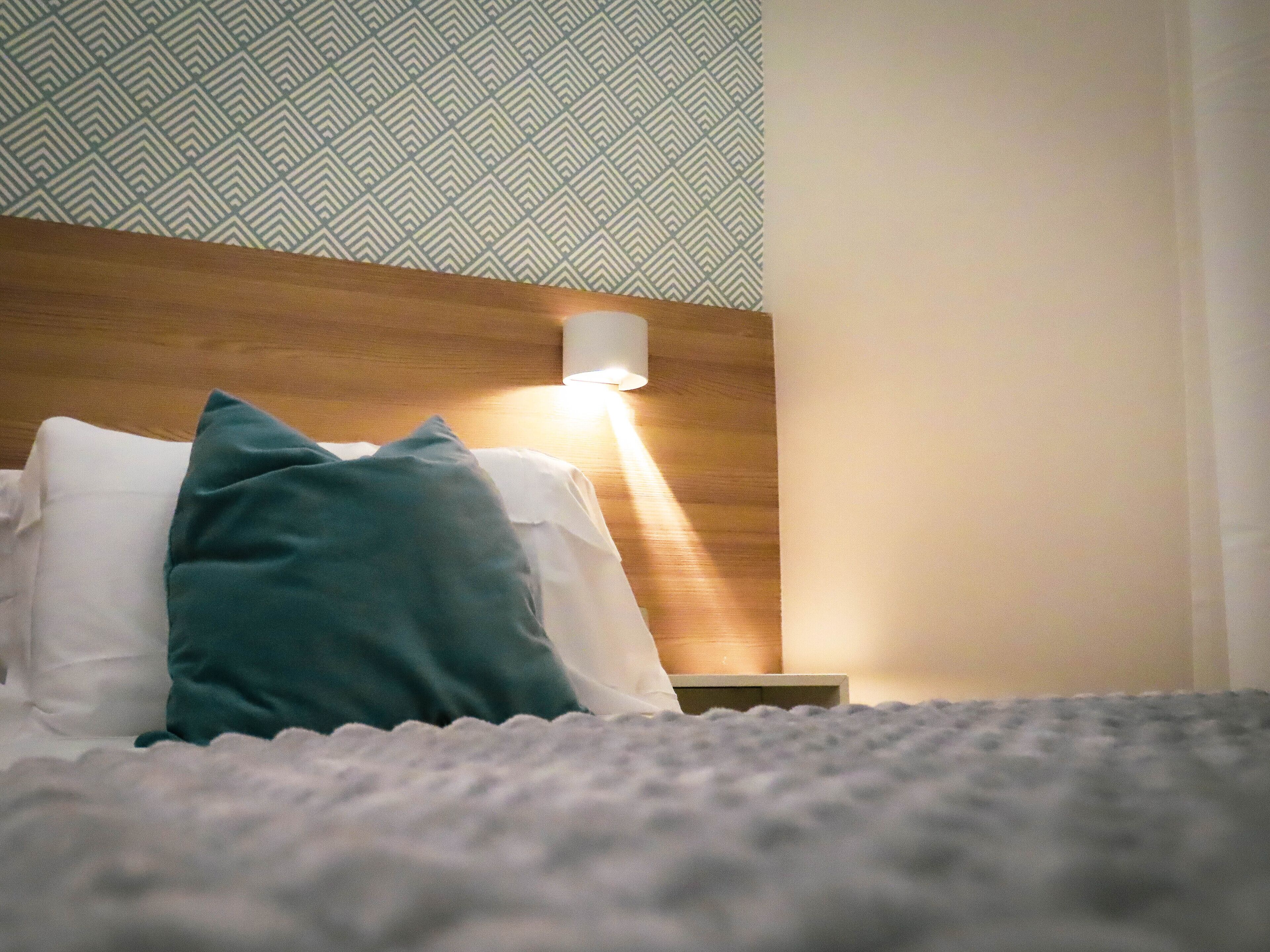 A neatly made bed with white and teal pillows, a wooden headboard, and a wall with a geometric pattern.