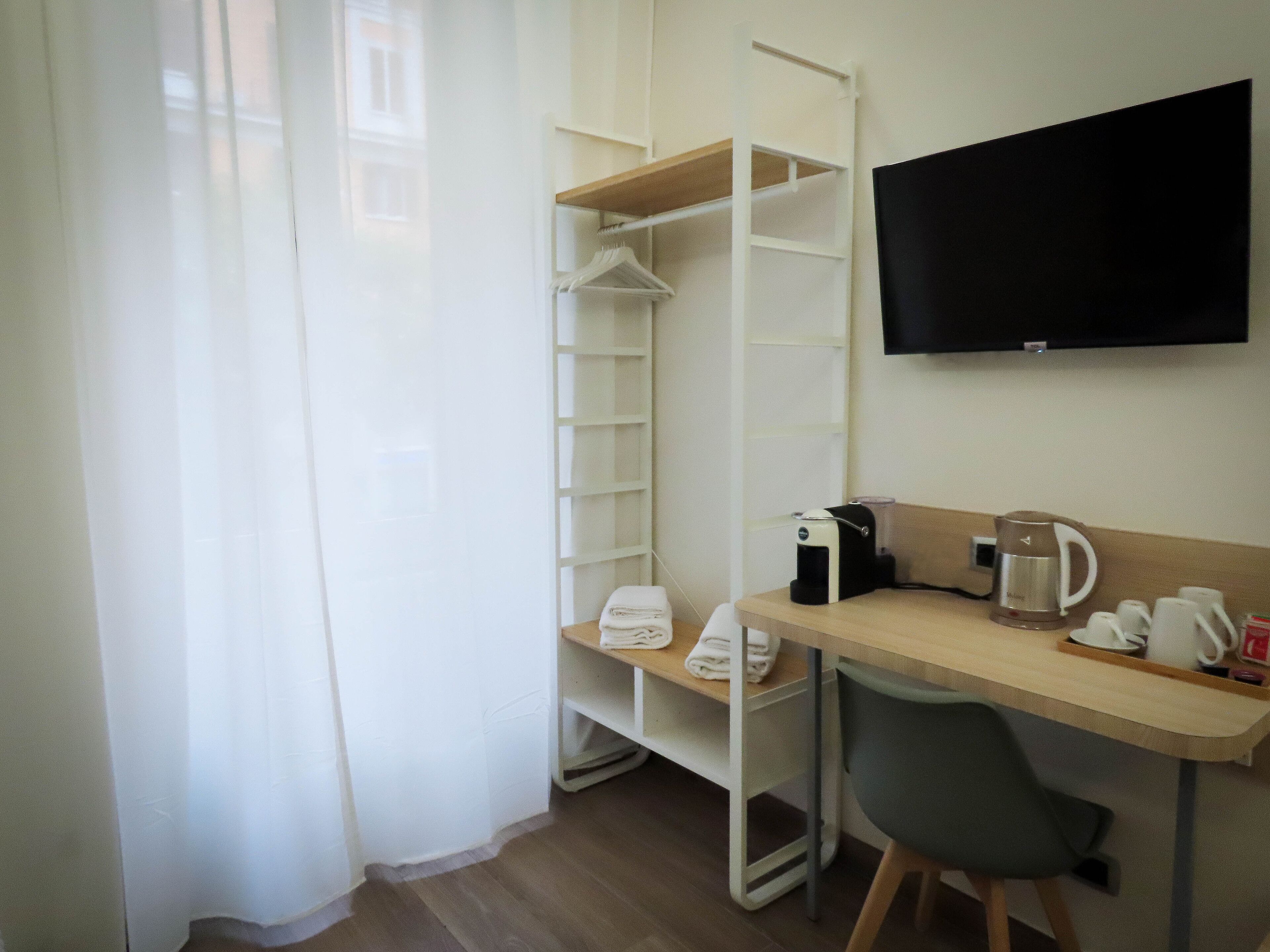 A compact hotel room with a wooden desk, a chair, a coffee maker, a TV, and a wardrobe with hanging clothes.