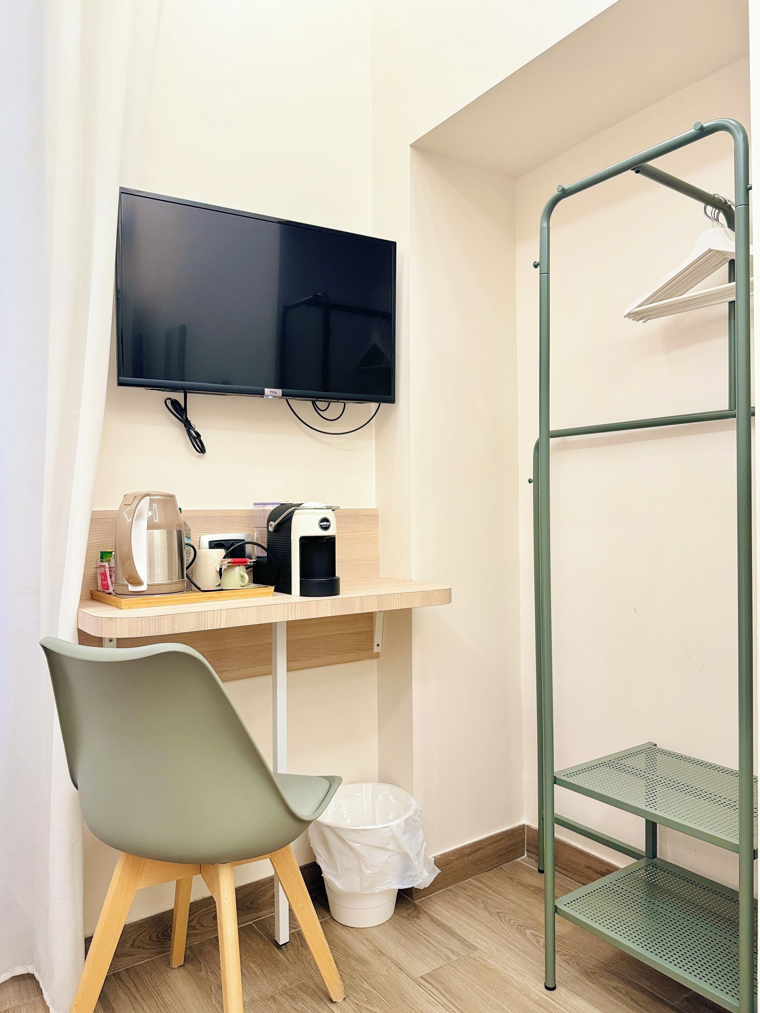 Ein kleiner, zweckmäßiger Raum mit Schreibtisch, Stuhl, Kaffeemaschine, Mülleimer und einem an der Wand befestigten Fernseher.