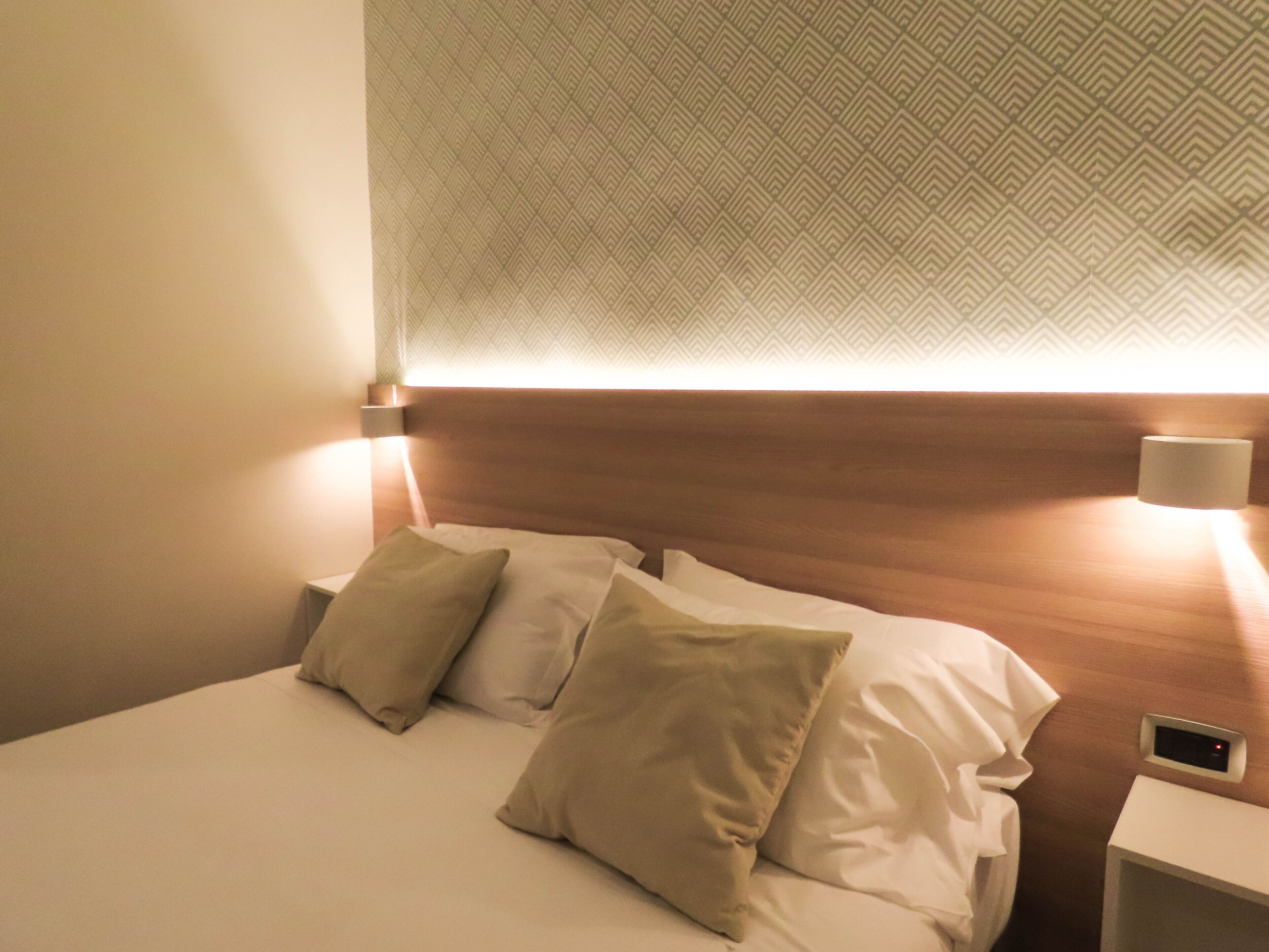 Una camera d'albergo con un letto rifatto, una testiera con carta da parati decorata e un comodino con una lampada.