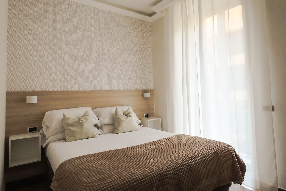 Ein Hotelzimmer mit einem großen Bett, zwei Nachttischen, einer Kopfteil und einem Fenster mit transparenten Vorhängen.
