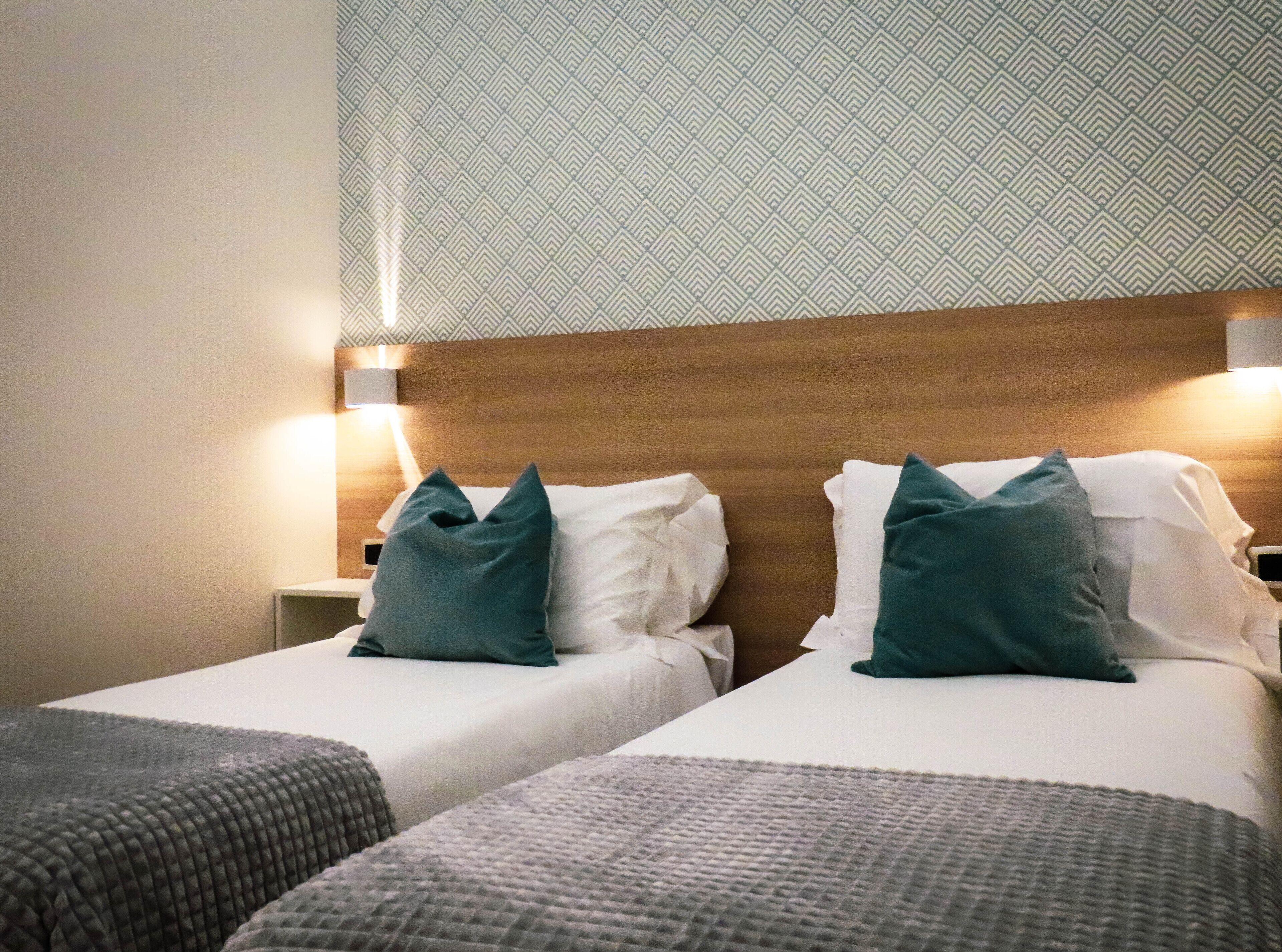 A hotel room with two single beds, a wooden headboard, a patterned wall, and a nightstand with a lamp.
