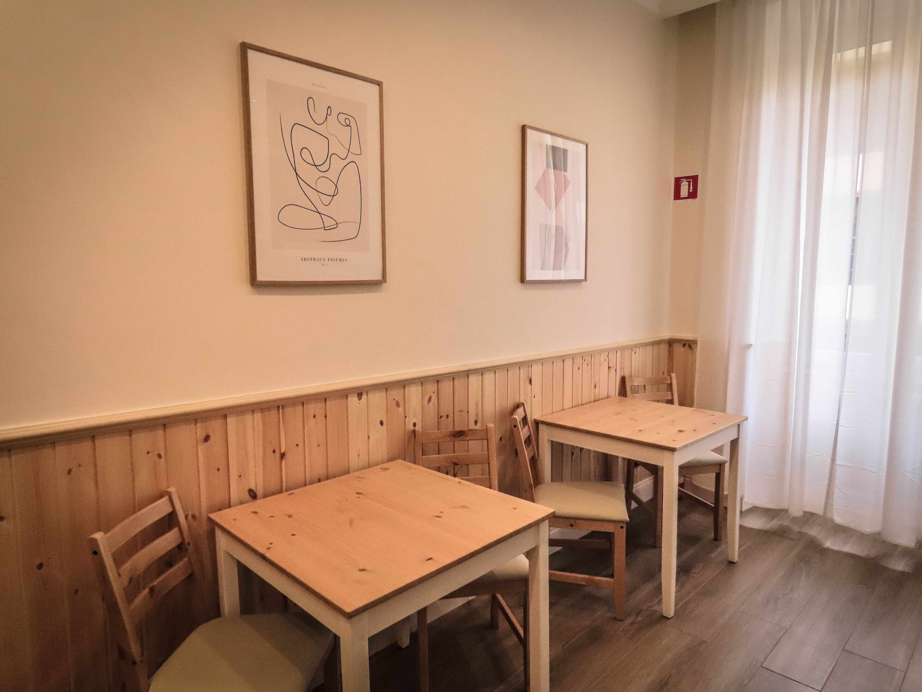 A room with two wooden tables and chairs, a wooden wainscoting, and two framed abstract artworks on the wall.