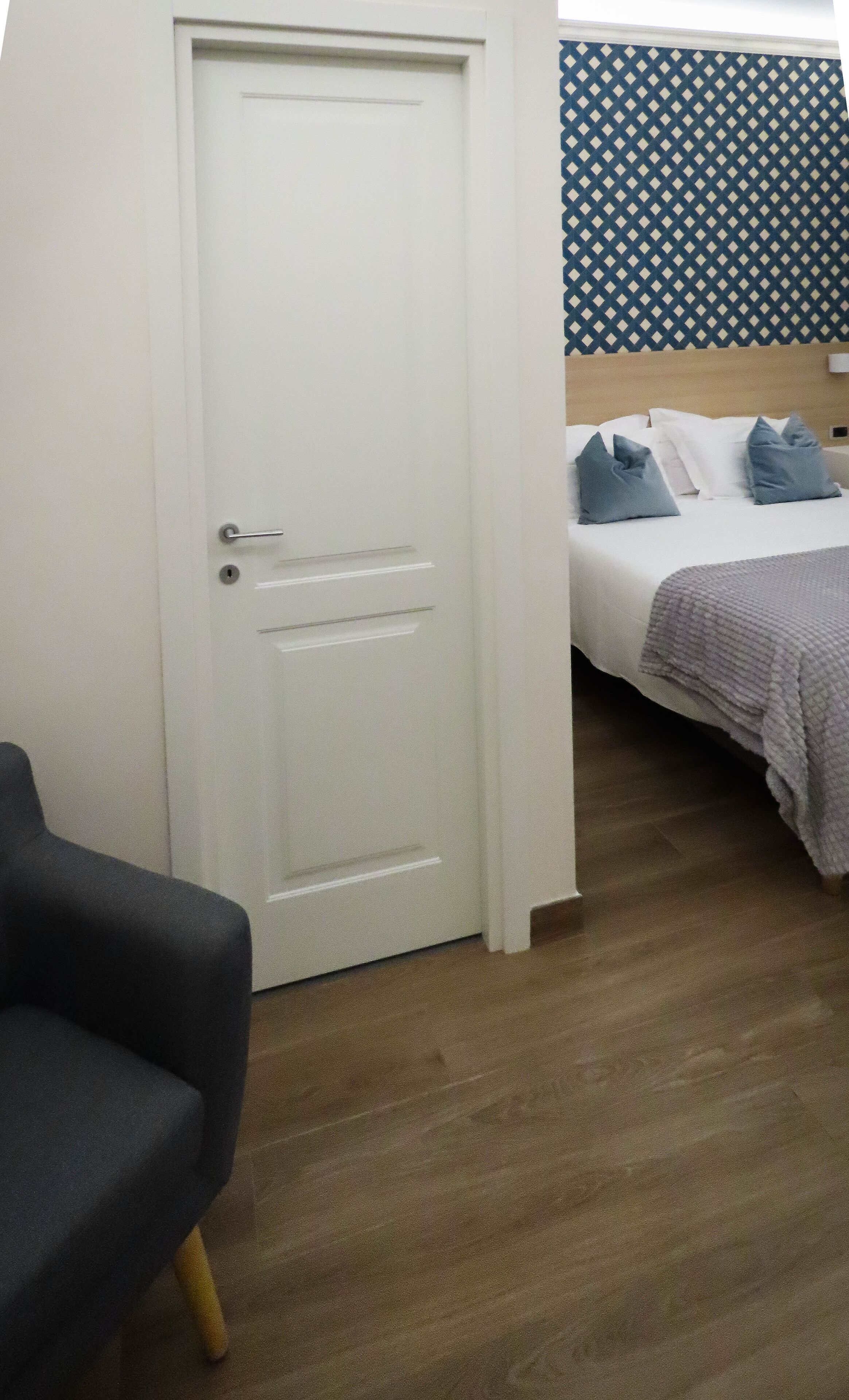 A hotel room with a white door, a bed with a headboard, a grey armchair, and a patterned wall.