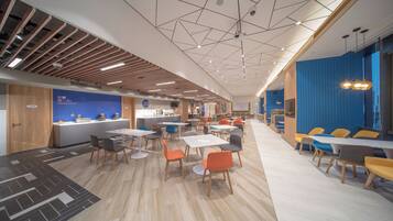 A modern cafeteria with multiple tables and chairs, a kitchen area, and a blue accent wall.