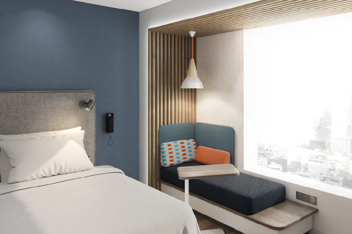 A modern hotel room with a bed, a seating area with a view of the city, and a cityscape visible through the window.
