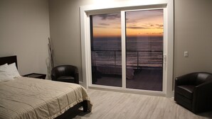 Un dormitorio con un amplio ventanal corredera que da a un balcón con vista al océano al atardecer.
