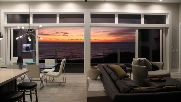 Una sala de estar moderna con un amplio ventanal que ofrece vista al atardecer sobre el océano, un ventilador de techo y muebles contemporáneos.