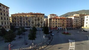 Une place avec une fontaine, entourée d’immeubles à plusieurs étages dotés de balcons, sous un ciel dégagé.